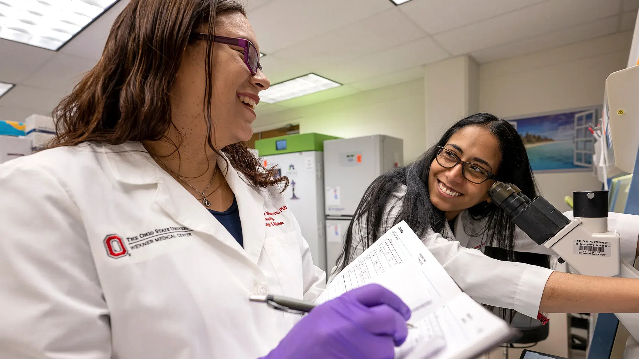 two women in ohio state lab coats discussing data in a lab next to microscopes